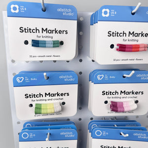 A retail display of All Stitch Studio stitch markers for knitting and crochet, showing multiple packages of different colors (blue, green, pink, yellow, purple) hanging on a white pegboard.