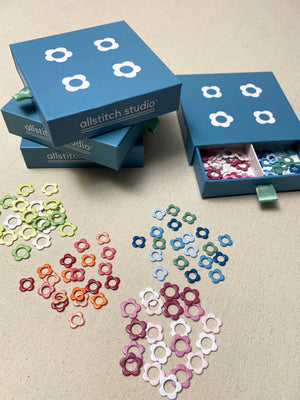 Several blue Allstitch Studio boxes are arranged on a beige fabric surface, with small flower-shaped ring embellishments scattered in front in multiple colors. One open box shows the interior compartments filled with the rings.