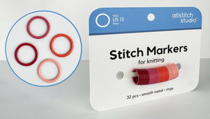 Allstitch Studio Stitch Markers for knitting, shown in their retail packaging with a blue header card. The product consists of 32 smooth metal rings in various colors including red, orange, and pink, which are displayed both inside the circular inset graphic and on the spool of the package.